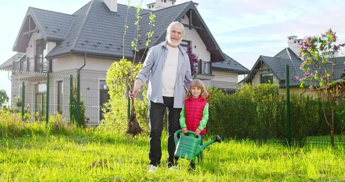 Portrait of happy young Caucasian grandfather with small tree with root in hand and little cute grandson with watering pot. Garden work in summer. Planting trees concept.