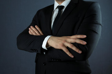 A man in a black suit on a dark background is standing in a half turn with his arms crossed close-up.