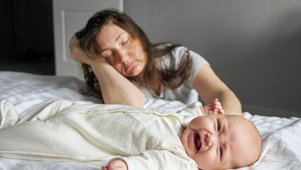 Tired mother sleeps sitting on floor leaning on bed with crying baby lying on bed white sheets....
