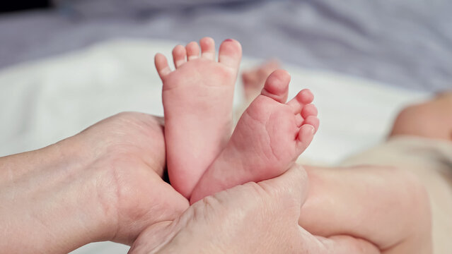 Woman holds bare feet of newborn baby making shape of heart on blurred background. Grandmother spends time with little child lying on bed closeup
