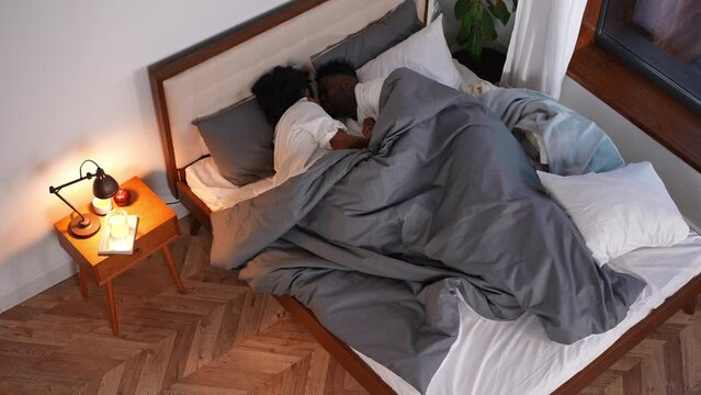 Wide shot loving African American couple cuddling lying in bed at home in the morning. High angle view portrait of smiling happy young man and woman rubbing noses resting in cozy bedroom indoors