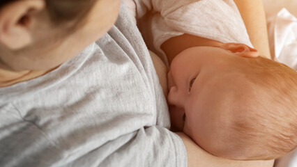 Mother feeds newborn baby with breast on bed at home. Adorable little girl in grey bodysuit sucks nipple lying in woman arms, close upper view