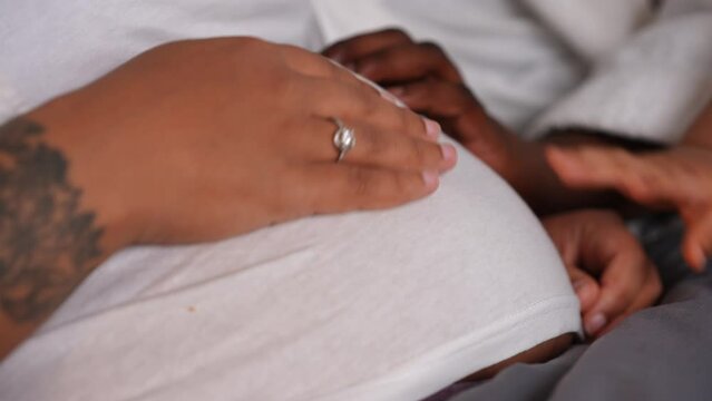 Close-up pregnant belly of African American young woman with hands caressing tummy. Unrecognizable husband wife and little son stroking belly lying in bed at home indoors. Family love concept