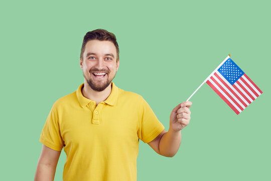 Happy young proud American celebrating Independence Day. Joyful white man having fun on July 4th. Studio portrait of cheerful handsome Caucasian guy holding flag of USA, looking at camera and smiling