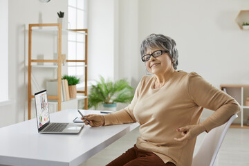 Fototapeta na wymiar Happy confident senior woman in glasses sitting at table with laptop computer at home, doing remote work, having video conference, studying business and finance courses. Remote work, education concept