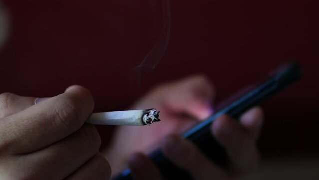 Man hand holding a cigar and typing on a smartphone screen in the blurred background in a dark room. The cigarette is smoking and the man is nervous
