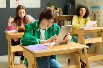 Fototapeta na wymiar Youthful female student in casualwear looking at tablet screen while scrolling through online hints at lesson among her classmates