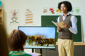 African American guy with copybook standing by interactive display with bees while making presentation in front of classmates at lesson