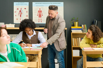 Mature male teacher of anatomy checking notes of highschool student and consulting him among classmates at lesson in classroom