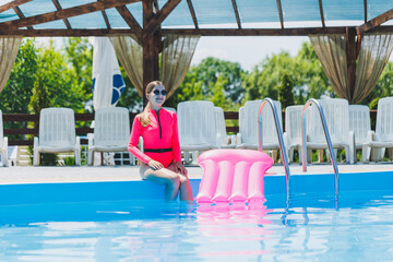 A beautiful woman in a pink swimsuit and with an inflatable mattress stands near a large pool. Summer holidays in a hotel with a swimming pool. Young girl in swimsuit and sunglasses