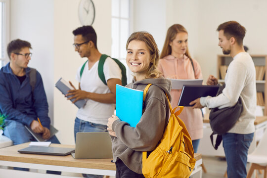 Portrait of happy smiling beautiful female university student with bag and notebook. Pretty college girl with candid cheerful face expression standing in classroom with her classmates in background
