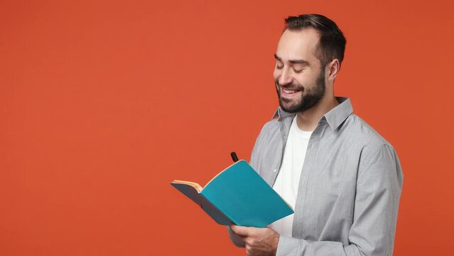 Happy fashionable swanky smiling young brunet man 20s years old wears blue shirt looking aside writing down in exercise book notebook diary notes isolated on plain orange background studio portrait