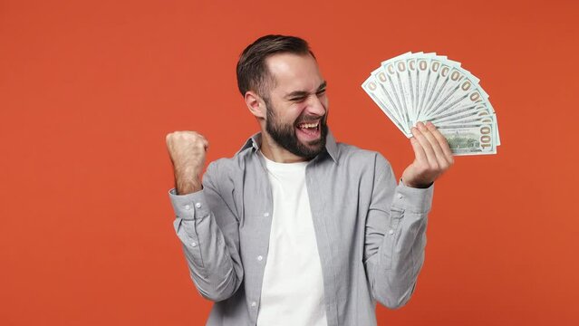 Young brunet man 20s years old wears blue shirt holding fan of cash money in dollar banknotes doing winner gesture celebrate clenching fists say yes isolated on plain orange background studio portrait