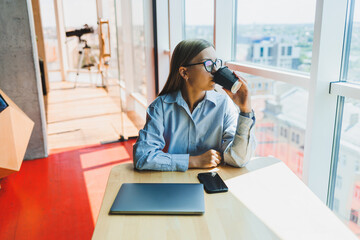 Successful woman is looking at a laptop in a cafe and drinking coffee. A young smiling woman in glasses sits at a table near the window with a phone. Freelance and remote work. Modern female lifestyle