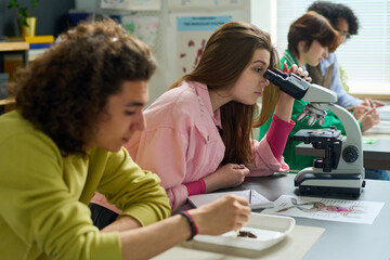 Teenage girl in casualwear studying dissected part of frog in microscope while sitting by desk next...