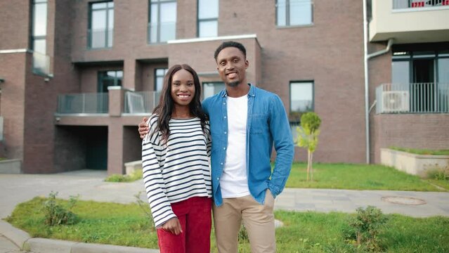 Portrait of happy young African American couple man and woman standing outdoors on street in front of residential building and smiling at camera. Love, family, happiness, relationship concept