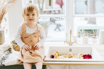 Fototapeta na wymiar cute little red haired girl is sitting in kitchen of country house with duckling bathing in sink, summer vibes concept, spring season and Happy Easter, baby ducks swimming with flowers