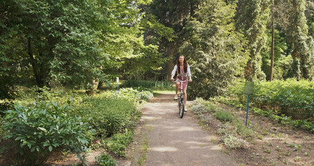 Obraz premium young woman rides a folding bike through a park with green trees towards the camera. The concept of a healthy lifestyle, eco movement, green transport. Wide shot, spring summer, daytime