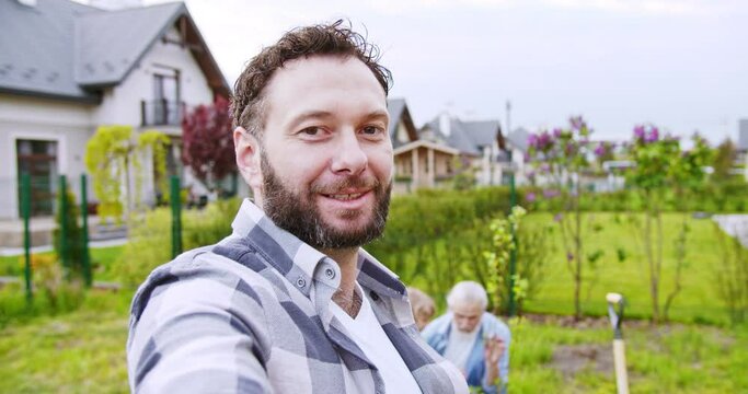 POV of handsome cheerful man taking selfie video or videochatting and showing old father with little cute son working in garden. Videochat. Summer work at house and planting.