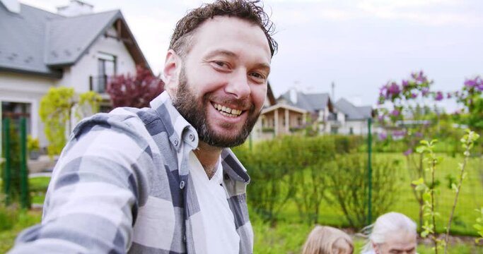 POV of happy man taking selfie video or videochatting and little cute son sending air kisses and waving with grandfather on the background. Working in garden. Videochat. Work at house and planting.