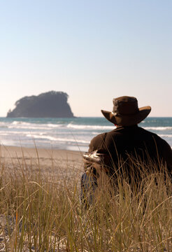 Back view of a man wearing an akubra hat and shoes slung over his shoulder sitting on the beach looking at the ocean