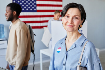 Waist up portrait of smiling adult woman at voting station smiling at camera with American flag in...
