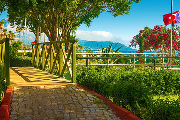 View of the path and the bridge in a green garden with green trees along the embankment in Kemer,...
