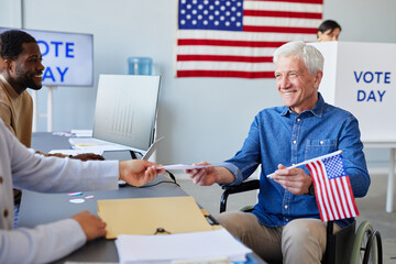 Portrait of smiling senior man in wheelchair receiving ballot at voting station on election day with USA flag in background
