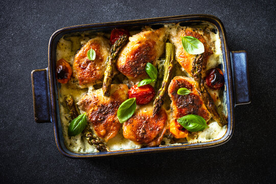 Baked chicken thighs with asparagus and tomatoes in a creamy sauce at black table.