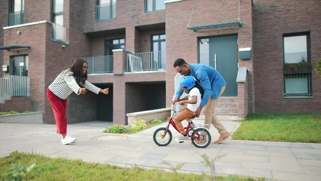 Happy family time together. Beautiful cheerful young mom and dad teaching little cute child boy how to ride a bicycle on street in city. Parents playing with son outdoor in neighbourhood area