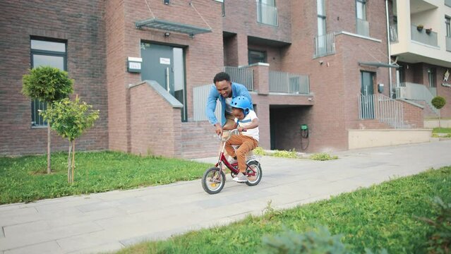 Joyful handsome African American young dad playing with little kid son teaching him how to ride a bicycle on street. Cheerful cute boy in helmet with father learning to ride a bike. Childhood concept