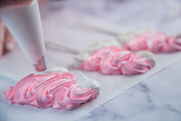 On light parchment there are pink meringue caps on sticks, the cook makes a pompom and a frill from a pastry bag of white meringue. Photography in a light key