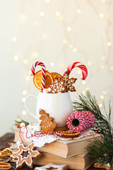 Hot winter drink in a white mug: cozy home composition with homemade gingerbread cookies, candy cane, fir tree branch. Wooden background, christmas lights and candles