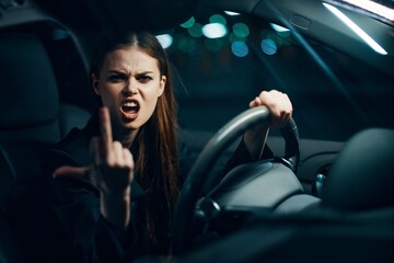 Fototapeta na wymiar an angry, aggressive woman sits behind the wheel of a car wearing a seat belt and shows a negative gesture to the camera emotionally screaming