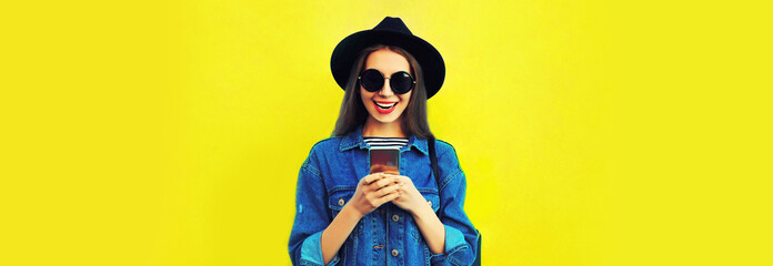 Obraz premium Portrait of stylish happy smiling young woman model with smartphone wearing black round hat, jean jacket on yellow background, blank copy space for advertising text