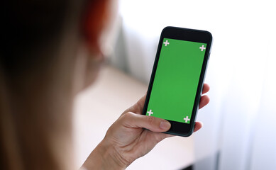 Chroma key mockup on smartphone in hand. Woman holds mobile phone iPhone and swipes photos or pictures left indoors of cozy home. Use green screen for copy space closeup.