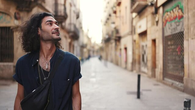 Young attractive italian guy with long curly hair and stubble looks at the architecture of the city. Stylish man with an earring in his ear and lot of chains. Tourist looking at the sights