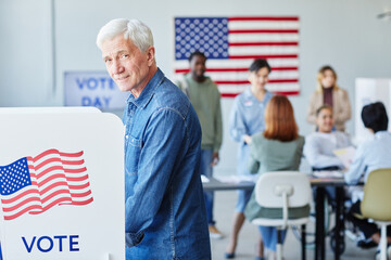 Side view portrait of smiling senior man voting in booth on election day and looking at camera, copy space