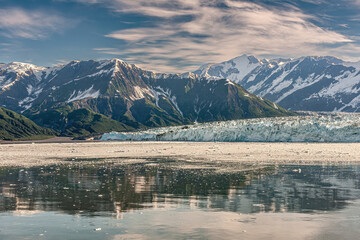 Disenchantment Bay, Alaska, USA - July 21, 2011: snow covered mountain range partly reflected in ocean water in front of Hubbard Glacier blue-white ice wall under blue cloudscape