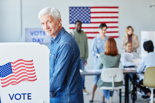 Side view portrait of smiling senior man voting in booth on election day and looking at camera, copy space