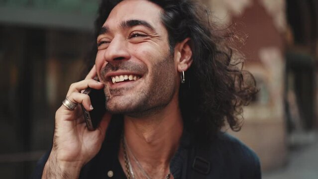 Clouse-up, attractive italian guy with long curly hair and stubble is using mobile phone at old buildings background. Stylish man with an earring in his ear and lot of chains talking on smartphone