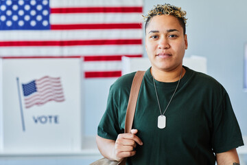 Waist up portrait of modern black woman looking at camera in voting station against American flag,...