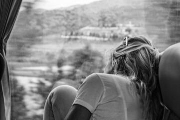 A black and white picture of a caucasian woman with long blond hair sitting in a bus staring outside the window of the landscape passing by with her raised knees on the bench.