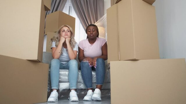Sad moving. Changing home. Changing life stress. Two tired upset depressed multiethnic female friends sighing on couch leaving apartment with isolated furniture unpacked boxes.