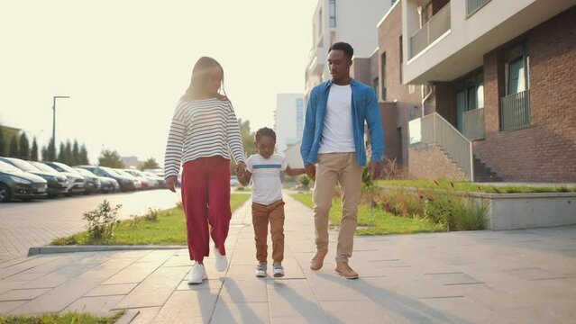 African American couple mom and dad walking with cute son on street in neighbourhood and smiling. Happy kid running toward camera. Family walk in suburb, spending time together, childhood concept