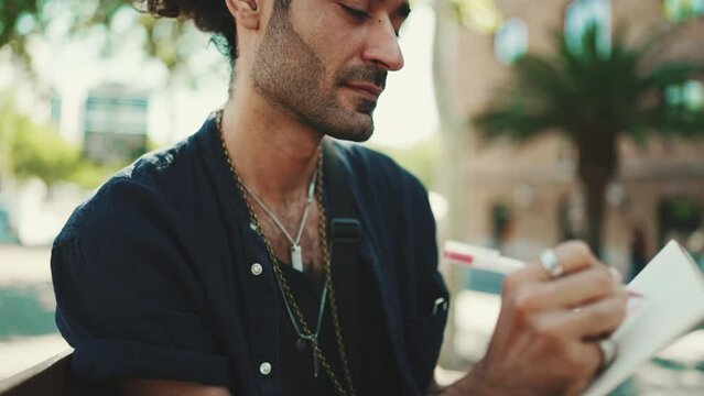 Close-up, young italian guy with ponytail and stubble sits on street bench and makes sketches with pen on sheet of paper on cityscape background