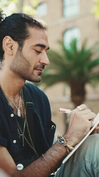 VERTICAL VIDEO: Young italian guy with ponytail and stubble sits on street bench and makes sketches with pen on piece of paper