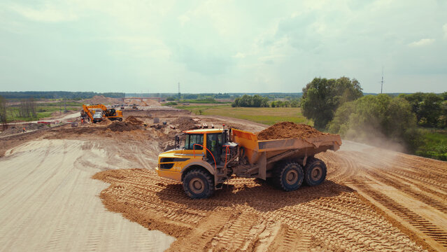 A yellow big dirty tipper truck filled up with a pile of sand standing at a road construction site. Other industrial machines in the background. High quality photo