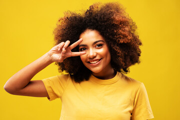young black curly woman 20s wears yellow shirt showing covering eye with victory sign