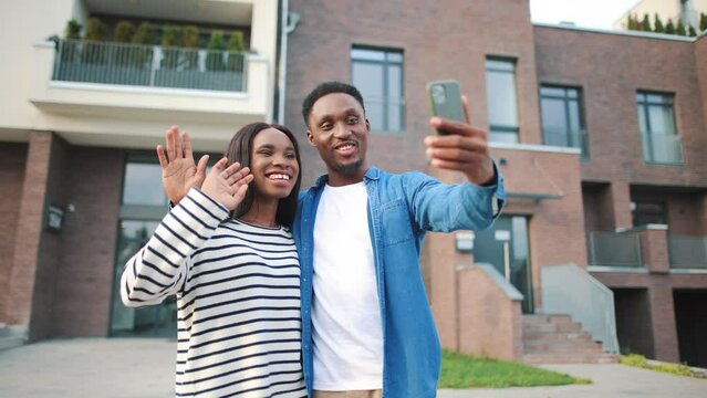 African American young cheerful family couple speaking on video call online on cellphone standing outside new home in neighbourhood and smiling. Happy man and woman video chatting on smartphone
